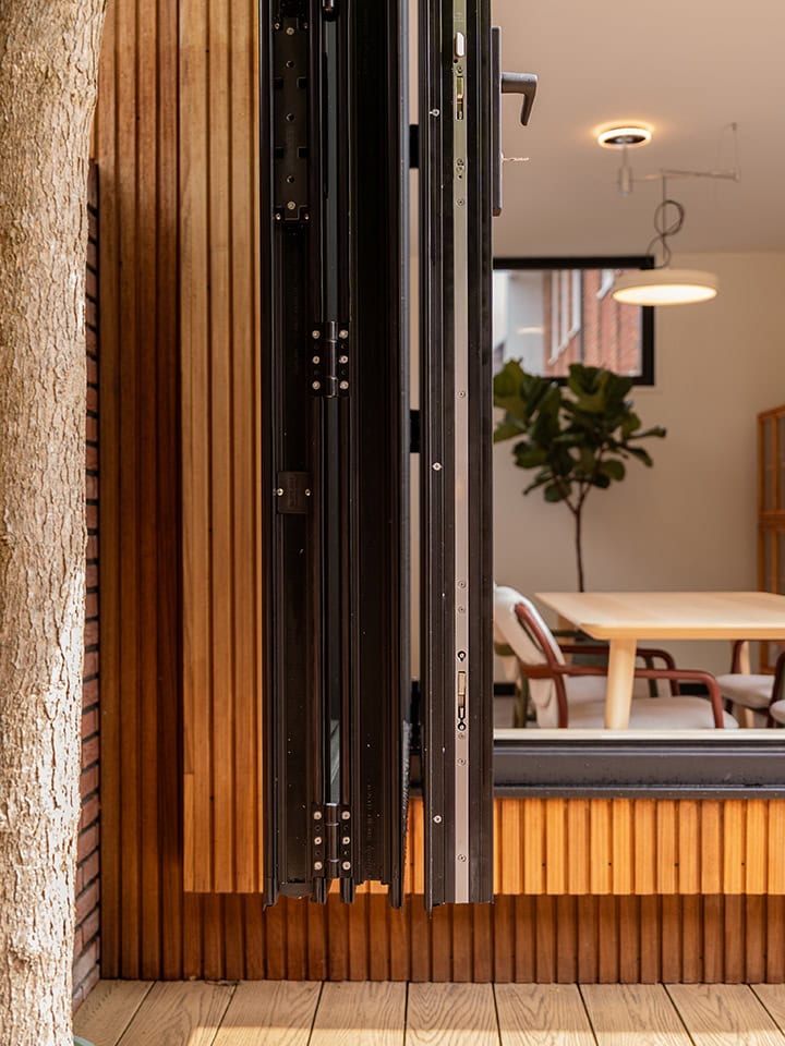 Close-up van een stalen deurmechanisme en frame, geïnstalleerd in een modern interieur met houten wandpanelen en een zithoek op de achtergrond.