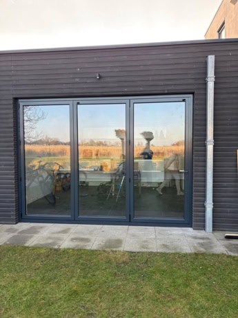 Moderne stalen schuifdeur met grote glazen panelen, perfect voor een strakke en eigentijdse uitstraling, geïnstalleerd aan de buitenkant van een zwart houten gebouw.