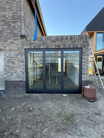 Moderne stalen schuifdeur met groot glasoppervlak, geïnstalleerd in een nieuwbouwwoning, met een bakstenen gevel en een heldere blauwe lucht op de achtergrond.