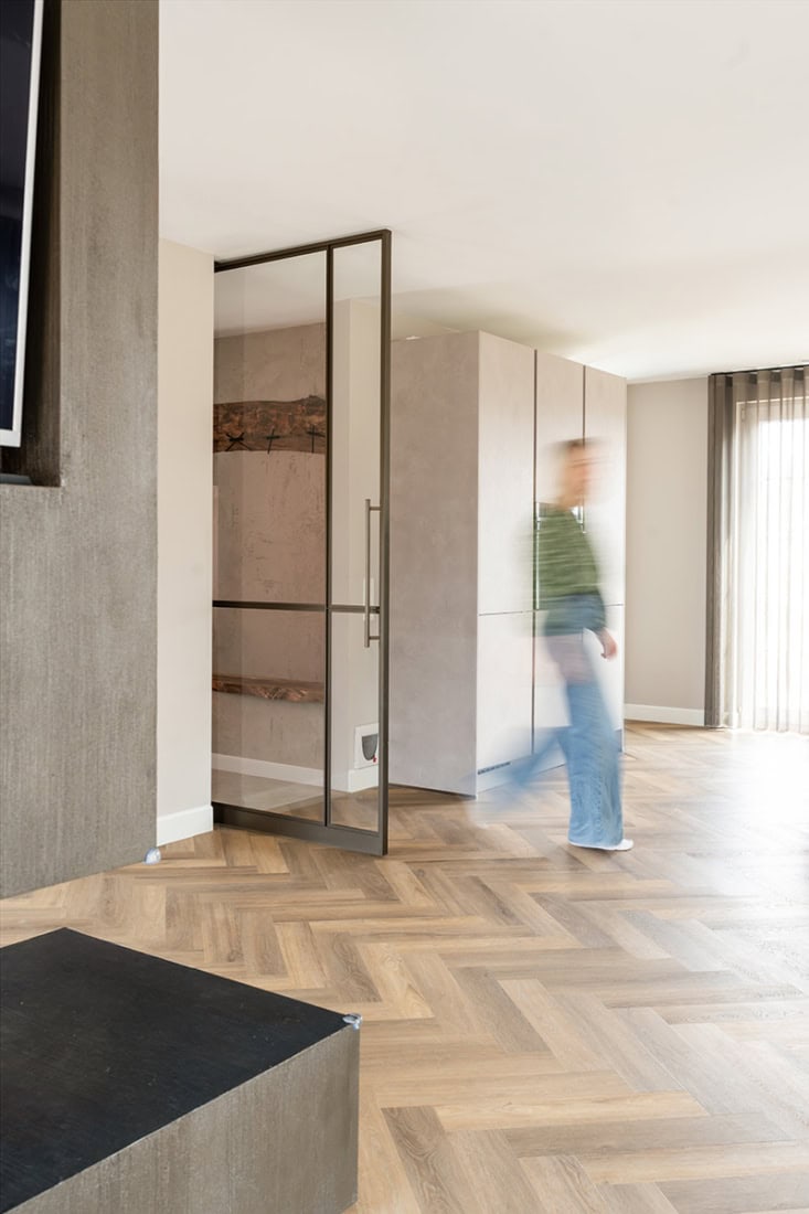 Stalen scheidingsdeur in moderne woonkamer met houten vloer en grote ramen, maatwerk staalwand voor strakke interieurafscheiding.