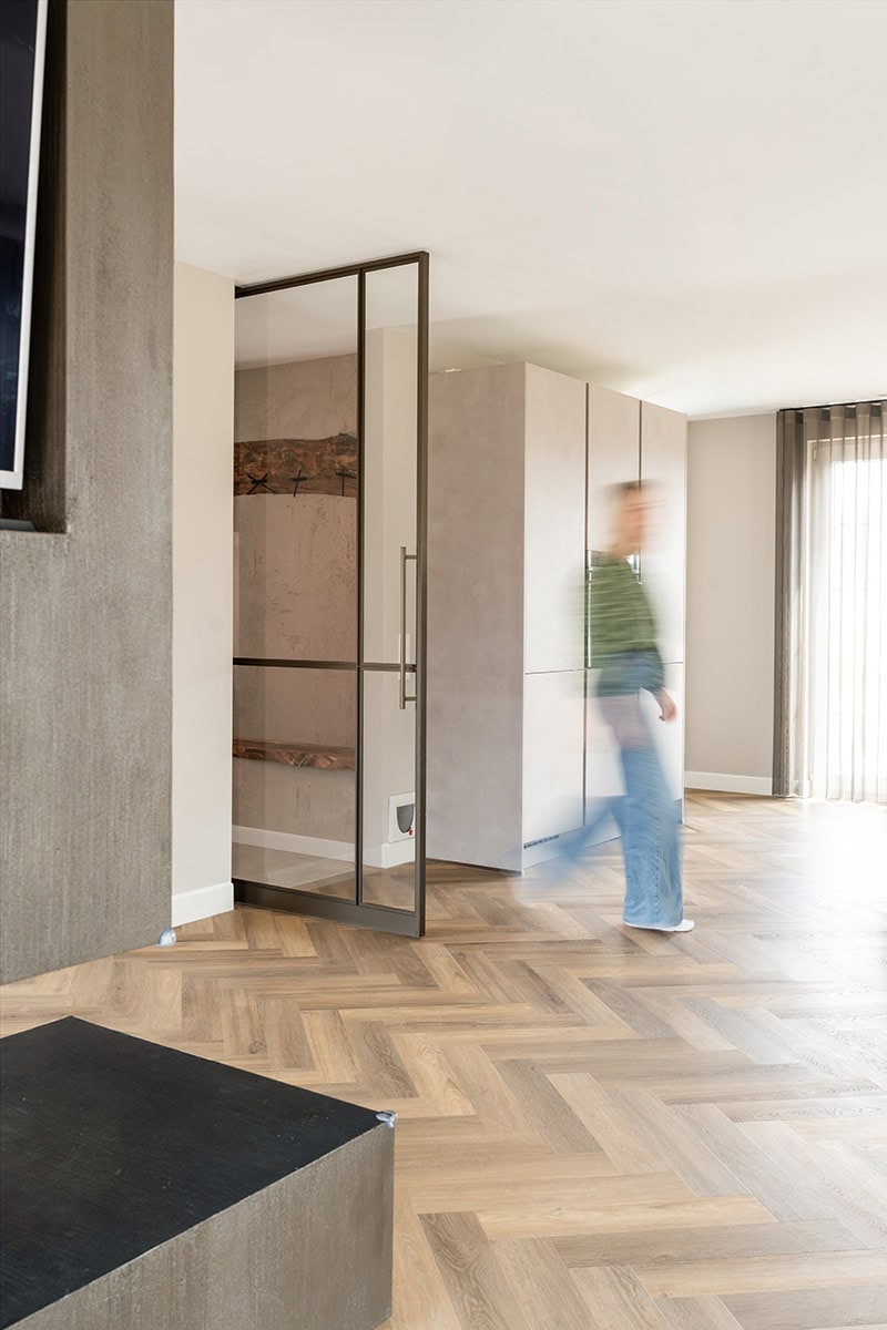 Stalen scheidingsdeur in moderne woonkamer met houten vloer en grote ramen, maatwerk staalwand voor strakke interieurafscheiding.