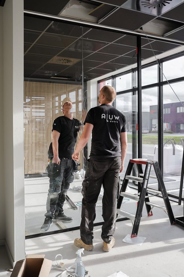 Glazen stalen deuropening met vakmanschap door Aluwdoors, montage in kantoorruimte, professionals werken aan op maat gemaakte industriële deuren, moderne bouwstijl, hoogwaardige afwerking, staal en glas.