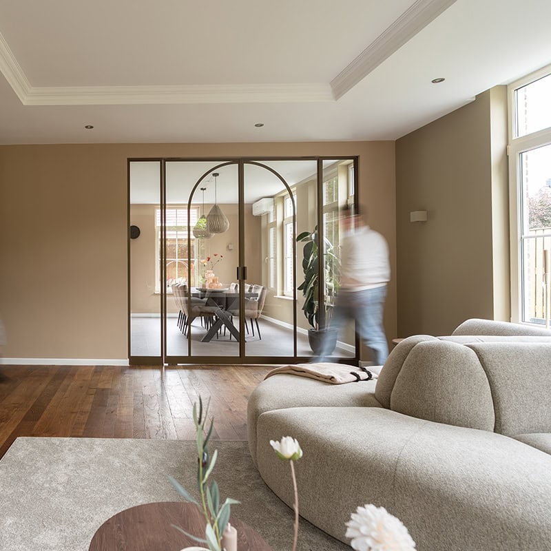 Sfeerbeeld van hoogwaardige stalen scheidingswand en deur op maat in moderne woonkamer met glazen schuifdeuren en natuurlijke lichtinval.