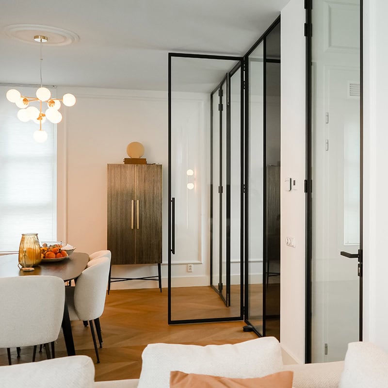 Rolgordijn van glas en zwart metalen kozijnen voor een moderne scheidingswand in een woonruimte.