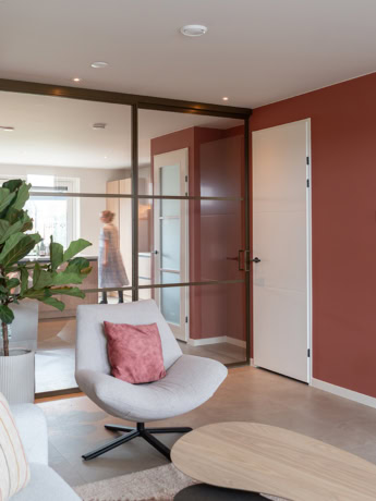 Moderne stalen deur en wand in een stijlvolle woonkamer, met een comfortabele stoel en een grote plant, passend bij de hoogwaardige stalen deuren en wanden op maat van Aluwdoors.